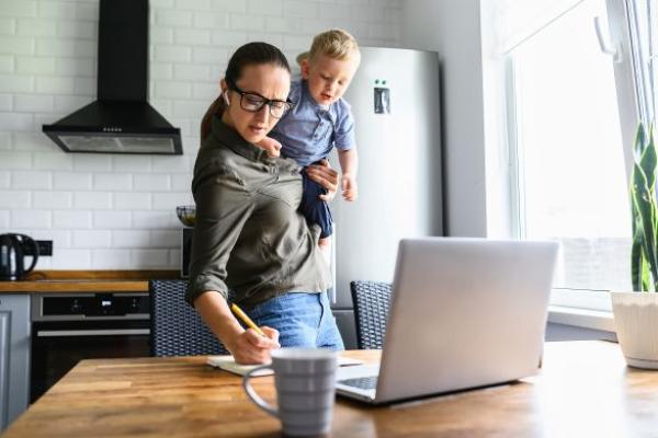 Maman débordée travaille à domicile avec son enfant actif dans les bras, prenant des notes tout en regardant son ordinateur portable.